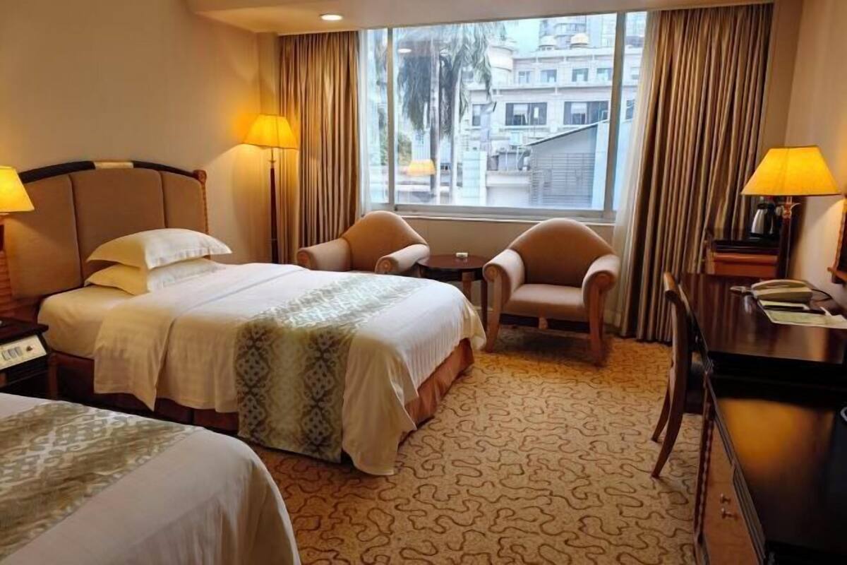 A hotel room with two beds, a desk, two chairs, a window with curtains, and a view of buildings outside.