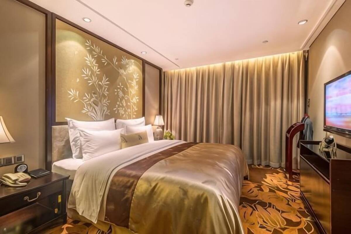 A hotel room with a bed, a TV, a desk with a lamp, and a large window with curtains.