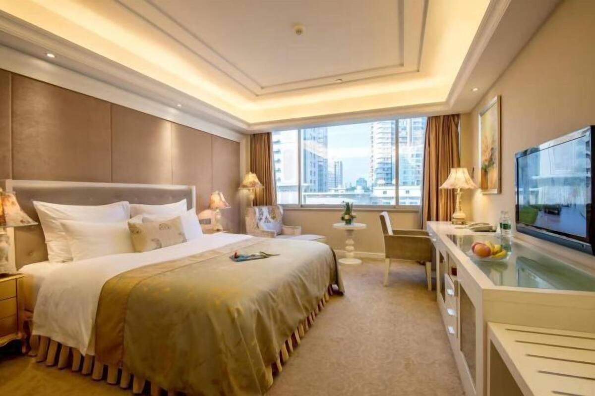 A hotel room with a large bed, a desk with a TV, and a window with a city view.