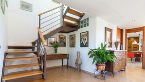 A spacious living area with a wooden staircase, a dining area with a red chair, and a kitchen visible in the background. - Villa es Baulo Pleta, with garden and pool near beach (Can Picafort)