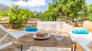Una mesa de madera con un plato de comida y un vaso, rodeada de sillones blancos y una toalla azul, ubicada sobre una zona arenosa con árboles y un muro de piedra al fondo.