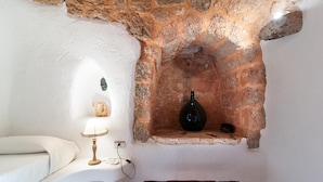 Una habitación rústica con pared de piedra, un banco de madera, una mesa blanca con una lámpara y una botella en un estante.