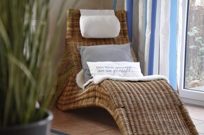 A wicker lounge chair with a white cushion and a pillow with text, positioned near a window with curtains.