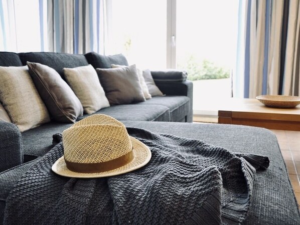 A cozy living room with a grey sofa, a woven hat on a blanket, and a view of the outdoors through large windows.