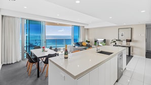 Quarto de hotel moderno com uma grande janela com vista para o mar, zona de refeições com uma mesa e cadeiras brancas, área de cozinha com pia e bancada e sala de estar com sofá e televisão.