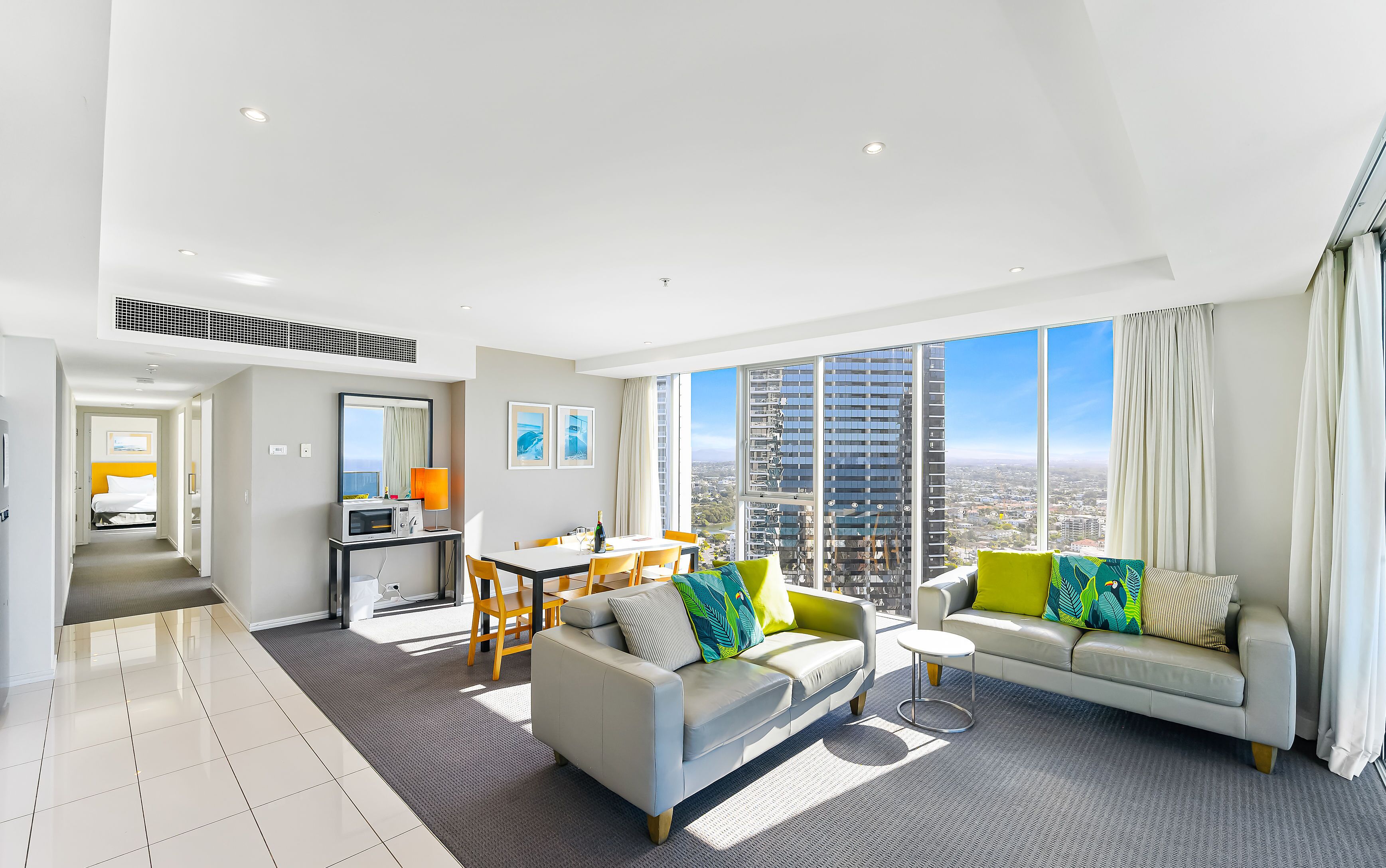 A modern hotel room with a dining area, a sofa set, and a large window offering a city view.