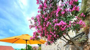 A stone patio with a table and chairs, a yellow umbrella, and a flowering tree. - Completely renovated small stone house with sea view near Split (Lokva Rogoznica)
