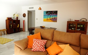Ein Wohnzimmer mit einem braunen Sofa, orangefarbenen Kissen, einem Esstisch und einem Gemälde mit Strandmotiv.