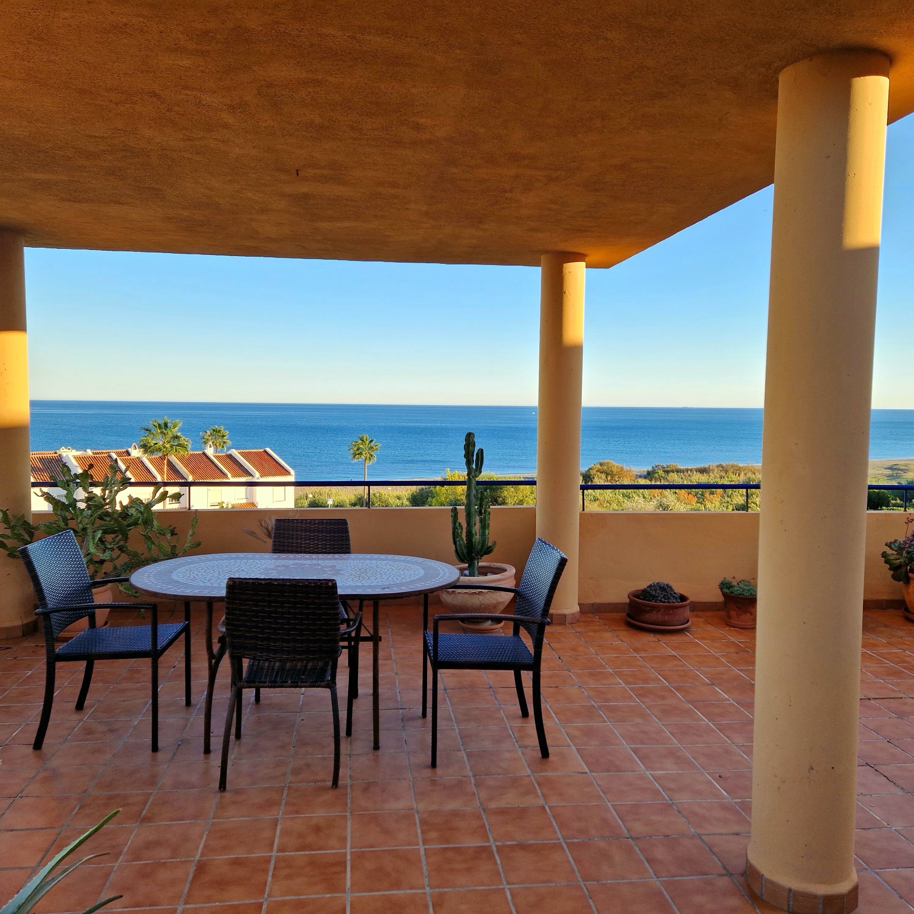 Um terraço com mesa e cadeiras redondas, um cacto e vista para o mar.