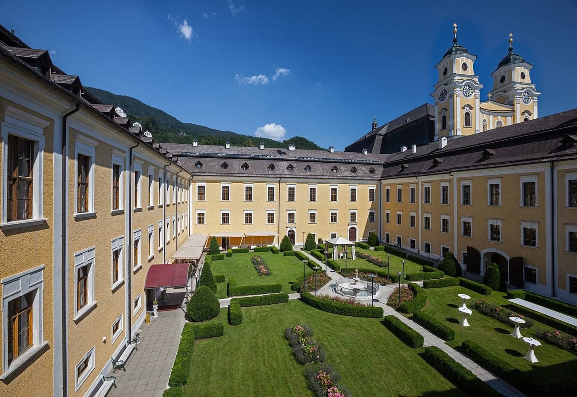 A courtyard with a central fountain, surrounded by manicured gardens and a building with two towers.