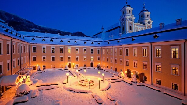 A snow-covered courtyard with a central fountain, surrounded by buildings with multiple windows and a clock tower in the background. - Live like a king - maisonette apartment in a castle hotel (Mondsee)