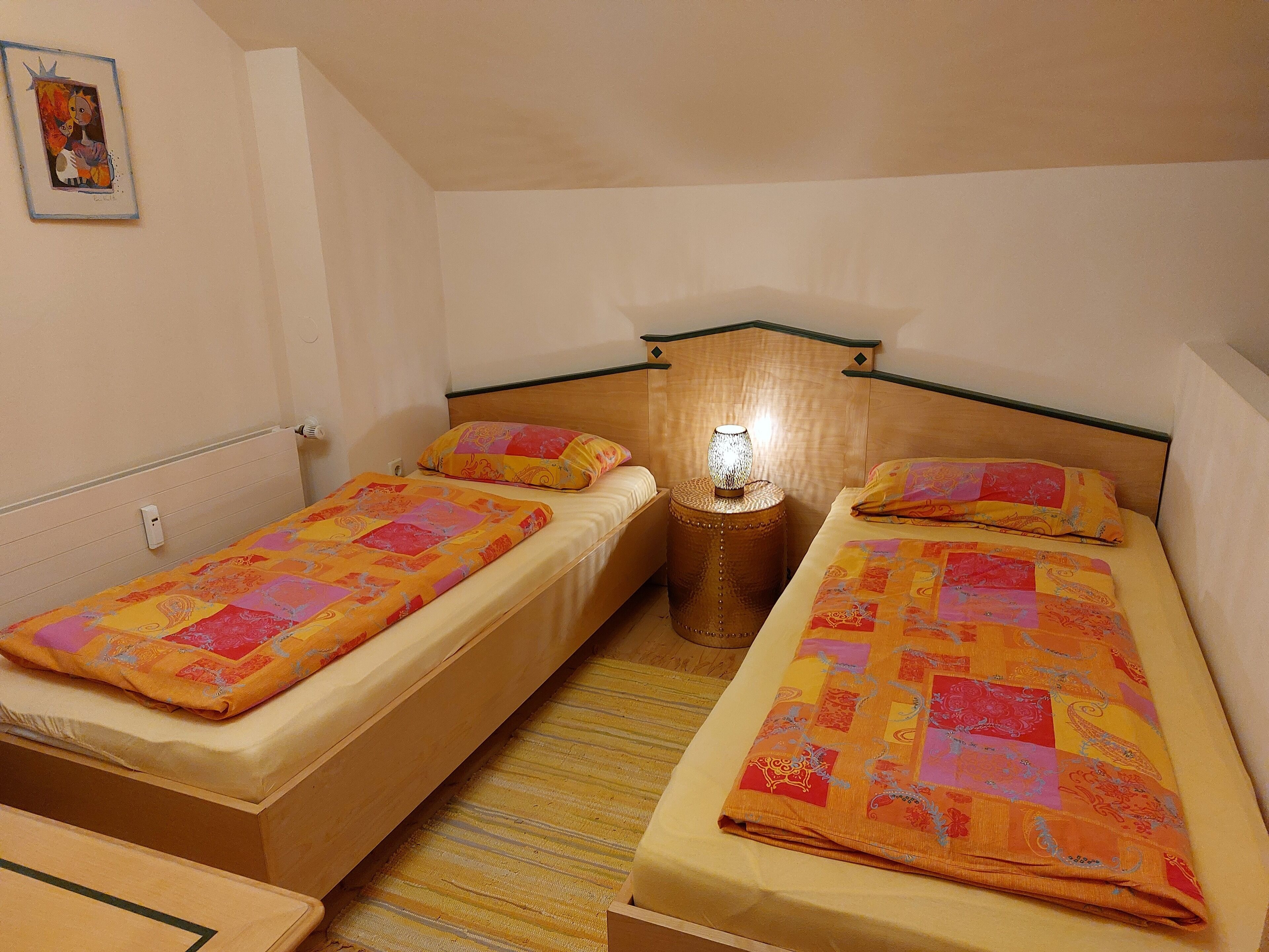 Ein kleines, gemütliches Zimmer mit zwei Einzelbetten, einem Nachttisch mit Lampe und einem gerahmten Bild an der Wand.
