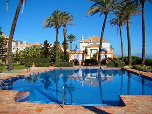 A swimming pool with clear blue water, surrounded by palm trees and a building with a tiled roof in the background. - Luxury apartment on ground floor with sea views, private garden (Bahia de Casares)