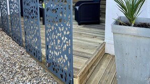 Uma área de estar moderna ao ar livre, com um deck de madeira, divisória de metal com desenhos recortados e um vaso de planta.