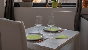 Uma sala de jantar com mesa posta para dois, uma janela com cortinas, uma planta em vaso e um objeto decorativo no parapeito.