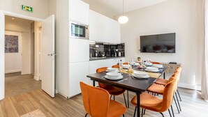 Een moderne keuken met witte kasten, een magnetron en een koffieapparaat. Er is een eettafel met oranje stoelen en een flatscreen televisie aan de muur bevestigd.