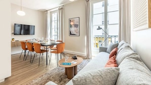 Een moderne woonkamer met een bank, eettafel en stoelen. Er is een televisie aan de muur, een raam met gordijnen, en een uitzicht over de stad.