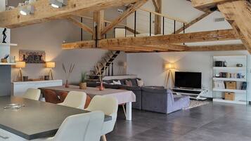 Een moderne loft met zichtbare houten balken, een eethoek, een bank, een televisie en een boekenkast.