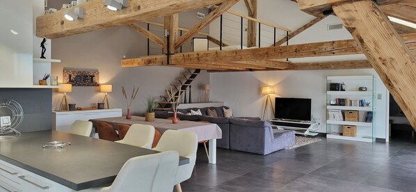 A modern loft-style living space with exposed wooden beams, a dining area, a sofa, a TV, and a bookshelf.