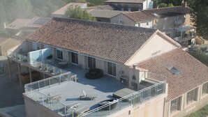 Un edificio de dos pisos con techo plano, un balcón con barandilla de vidrio y un patio con mobiliario exterior.