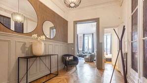 A hallway with a wooden floor, a console table with a vase of flowers, a round mirror, and a modern light fixture. - Sweett | Saint Lazare (Paris)