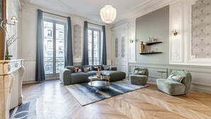 A spacious living room with a large sofa, a coffee table, and a fireplace. There are large windows with curtains, a chandelier, and decorative wall sconces. - Sweett | Saint Lazare (Paris)