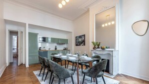 A dining area with a table set for six, surrounded by six chairs. A kitchen is visible in the background with modern appliances. - Sweett | Saint Lazare (Paris)