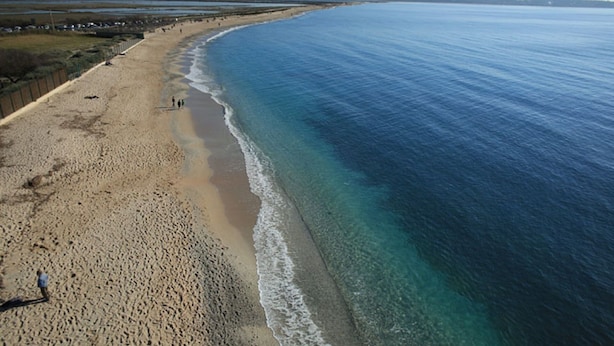 A wide beach with clear blue water, sandy shore, and a few people walking.