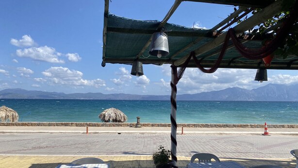 A seaside restaurant with outdoor seating, a view of the ocean, and mountains in the background.