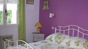 Een slaapkamer met een wit metalen bed, beddengoed met groene bloemen, een klein nachtkastje met een lamp en een raam met groene gordijnen.