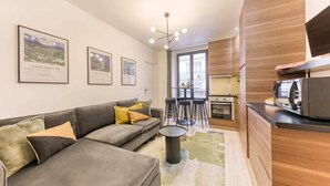 A modern living room with a grey sofa, a coffee table, and a kitchen area with wooden cabinets and a microwave. - Sweett | Troyon - Three Bedroom Apartment, Sleeps 6 (Paris)