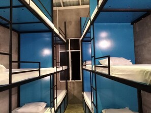 Habitación estrecha con literas dispuestas en un pasillo central, paredes azules y suelo de hormigón.