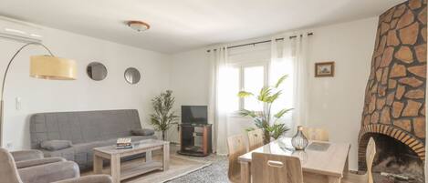 Una sala de estar con chimenea de piedra, un sofá gris, una mesa de centro y una mesa de comedor con sillas.