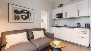 A modern living room with a sofa and a coffee table, a kitchenette with white cabinets and a microwave, and a framed abstract artwork on the wall. - Sweett | Monsigny V - One Bedroom Apartment, Sleeps 3 (Paris)