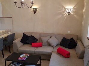Un salon avec un canapé beige, deux coussins noirs et un coussin rouge, une table basse noire sur laquelle sont posés des magazines, et des appliques murales.