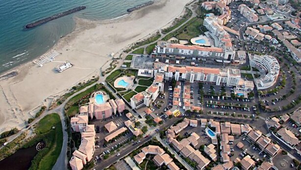 Oversigtsbillede af et kystresort med en strand, swimmingpools og forskellige bygninger, herunder et hotel med en buet facade og en central gårdsplads.
