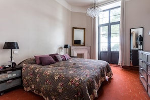 Une chambre à coucher avec une couvre-lit à motifs floraux, une cheminée, une grande fenêtre et une commode.