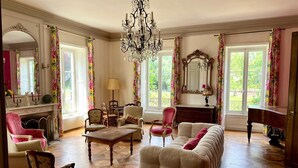 Een ruime woonkamer met een grote bank, een houten salontafel en een piano. Er staan verschillende stoelen en er is een open haard. De kamer heeft grote ramen met bloemen gordijnen en een kroonluchter.