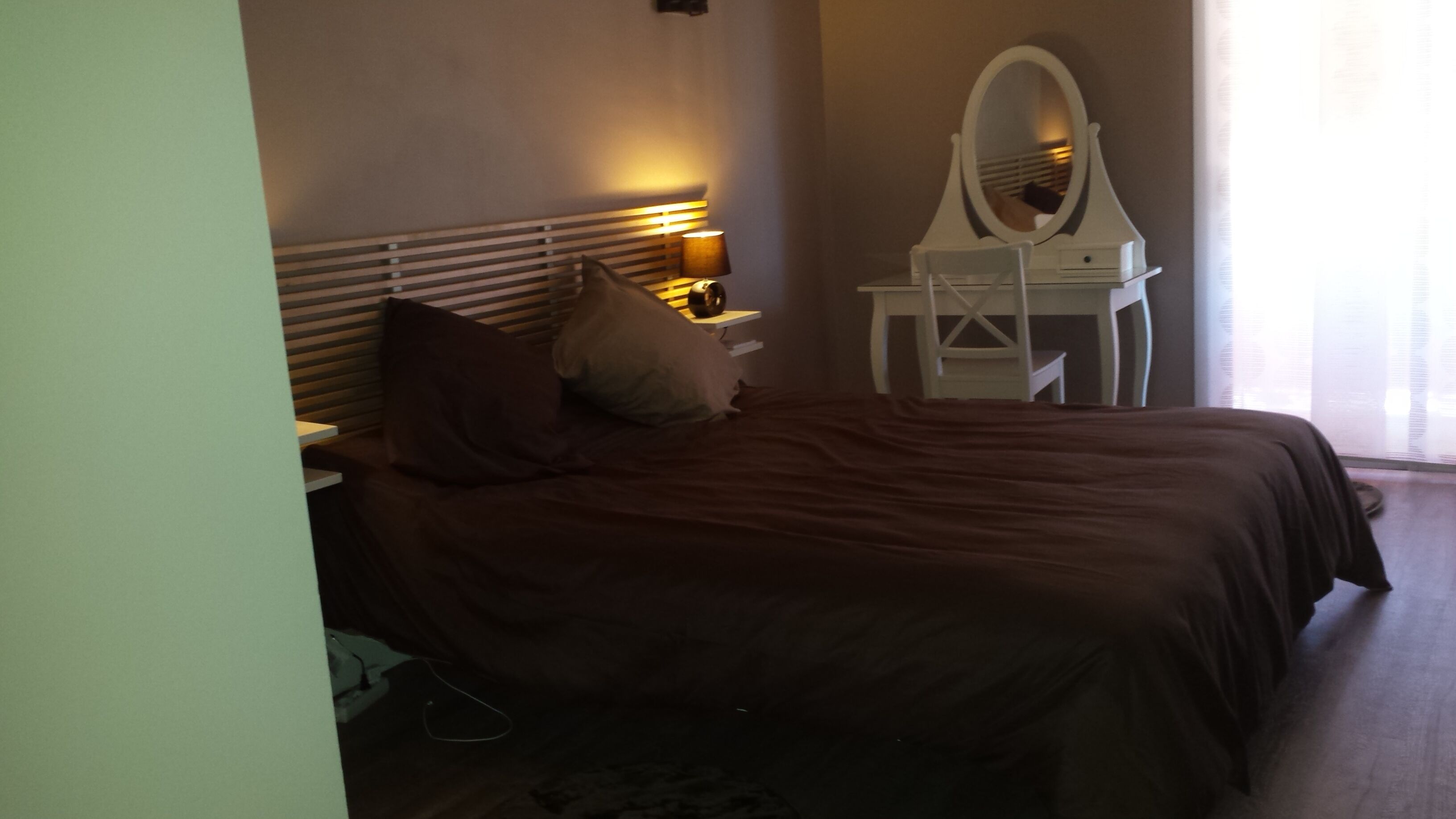 A bedroom with a bed, a bedside table, a lamp, a mirror, and a window.