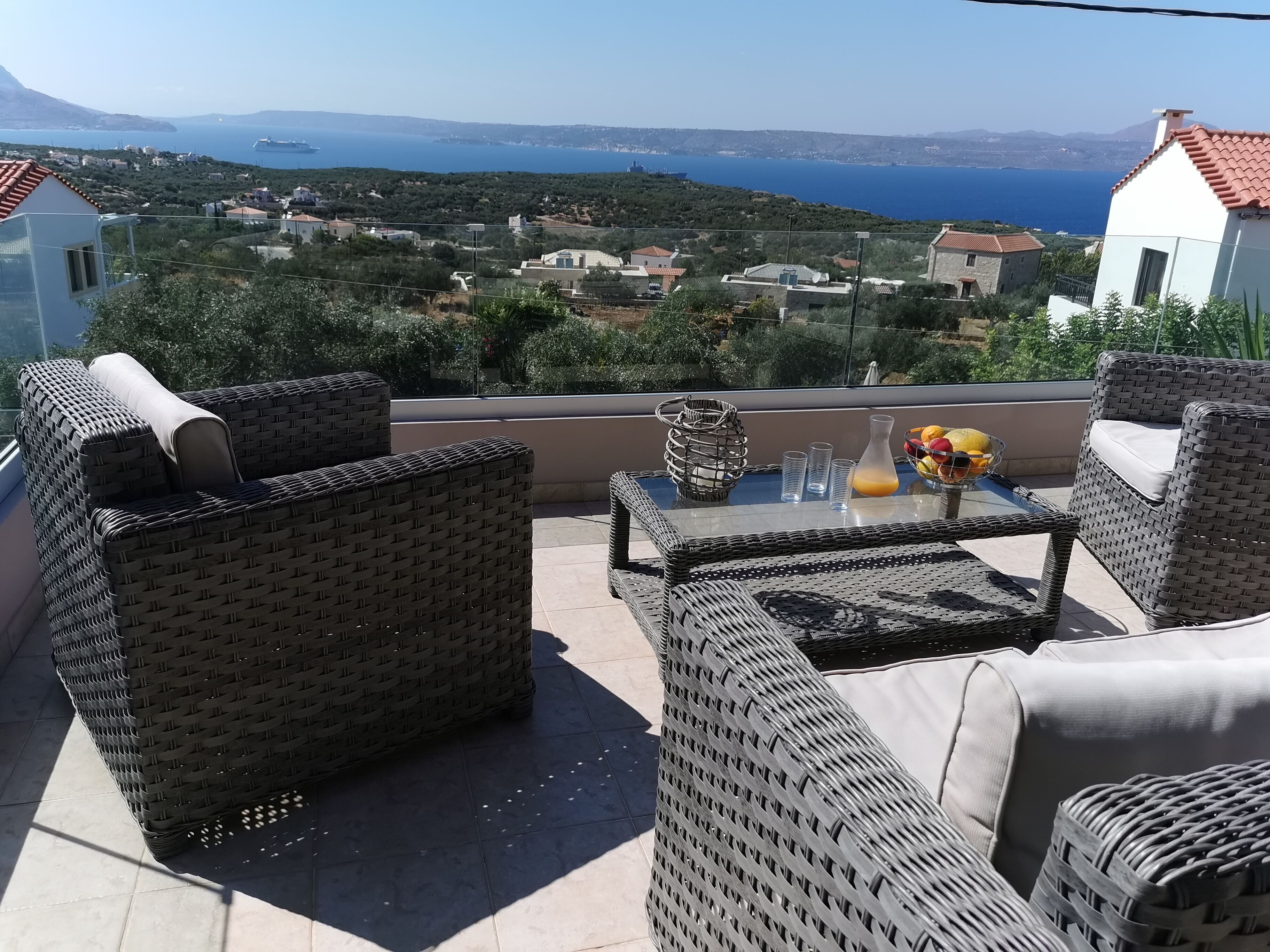 A terrace with wicker furniture, a glass table, and a view of the sea and hills.