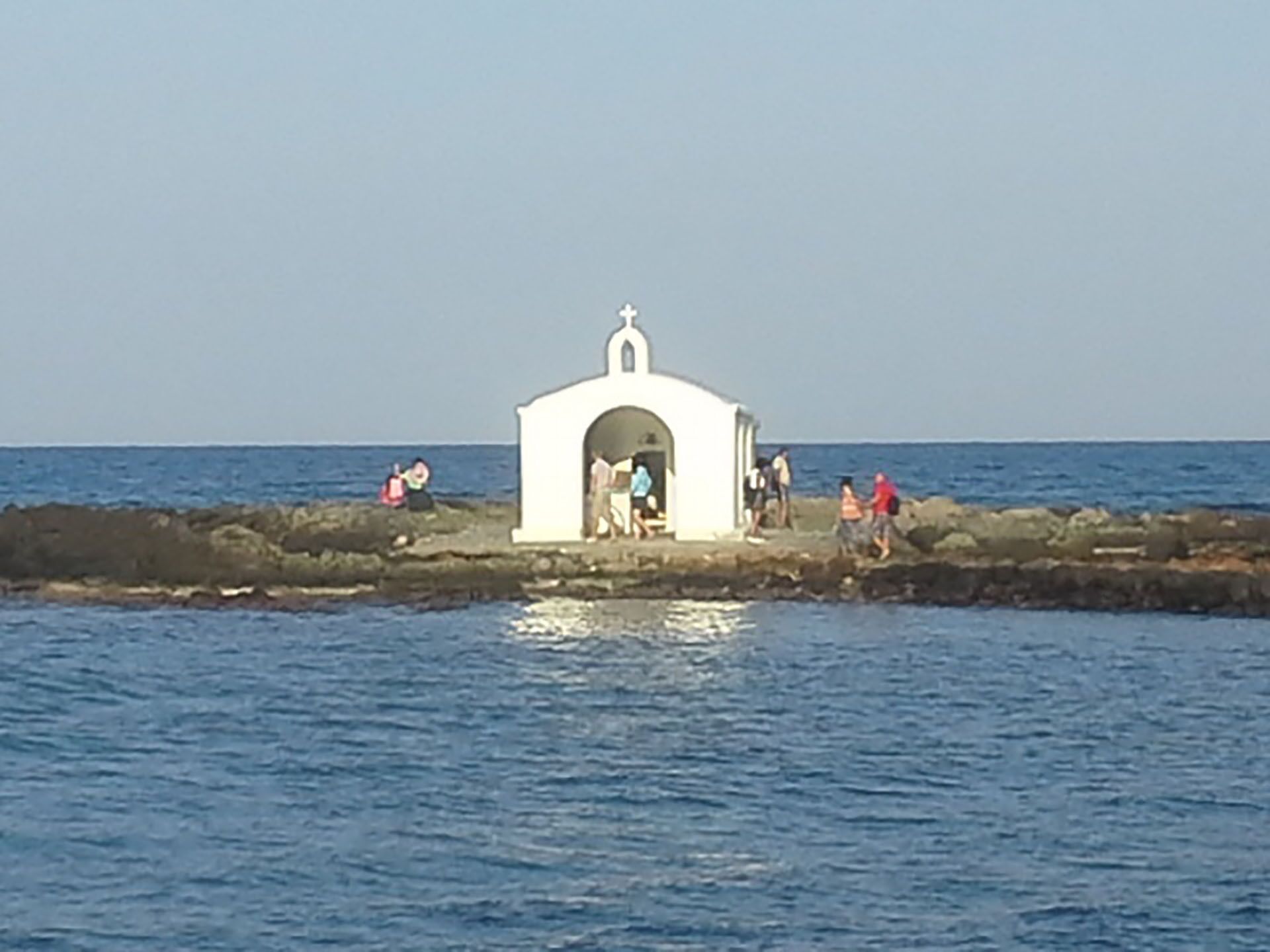 A small white building with a cross on top, located on a rocky shoreline by the sea.