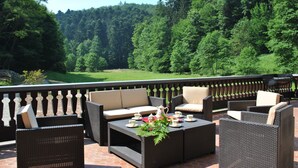 An outdoor patio with wicker furniture, a coffee table with a floral arrangement, and a scenic view of a forested area.