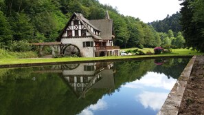 A traditional half-timbered house with a large water wheel, surrounded by lush greenery and a calm pond reflecting the structure. - A holiday in the Petite Pierre in the middle of the forest (Lohr)