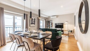 A modern dining area with a wooden table, chairs, and pendant lights. There is a green sofa and a TV in the background.
