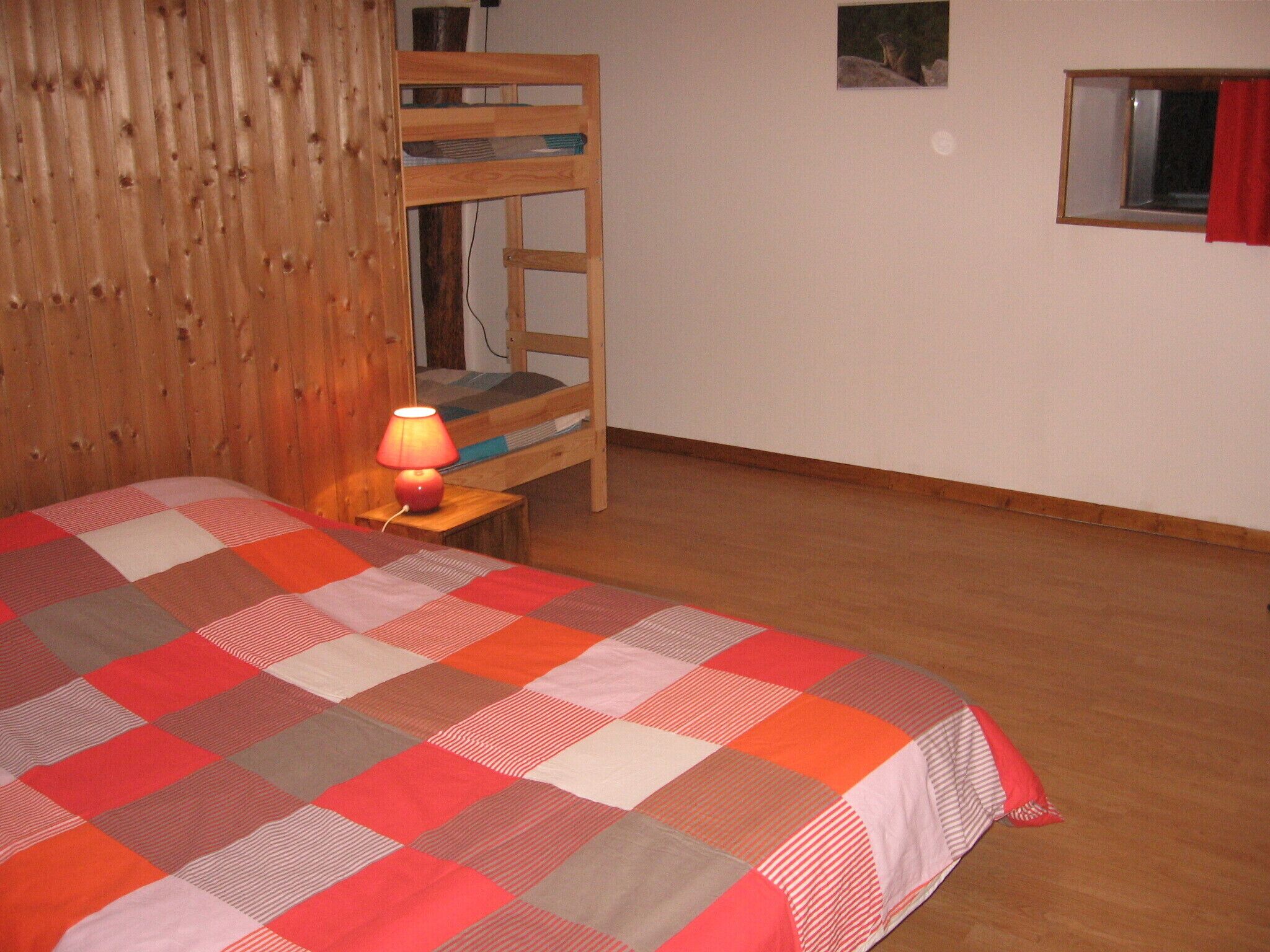 A bedroom with a wooden bunk bed, a red and white checkered bedspread, and a small bedside table with a lamp.