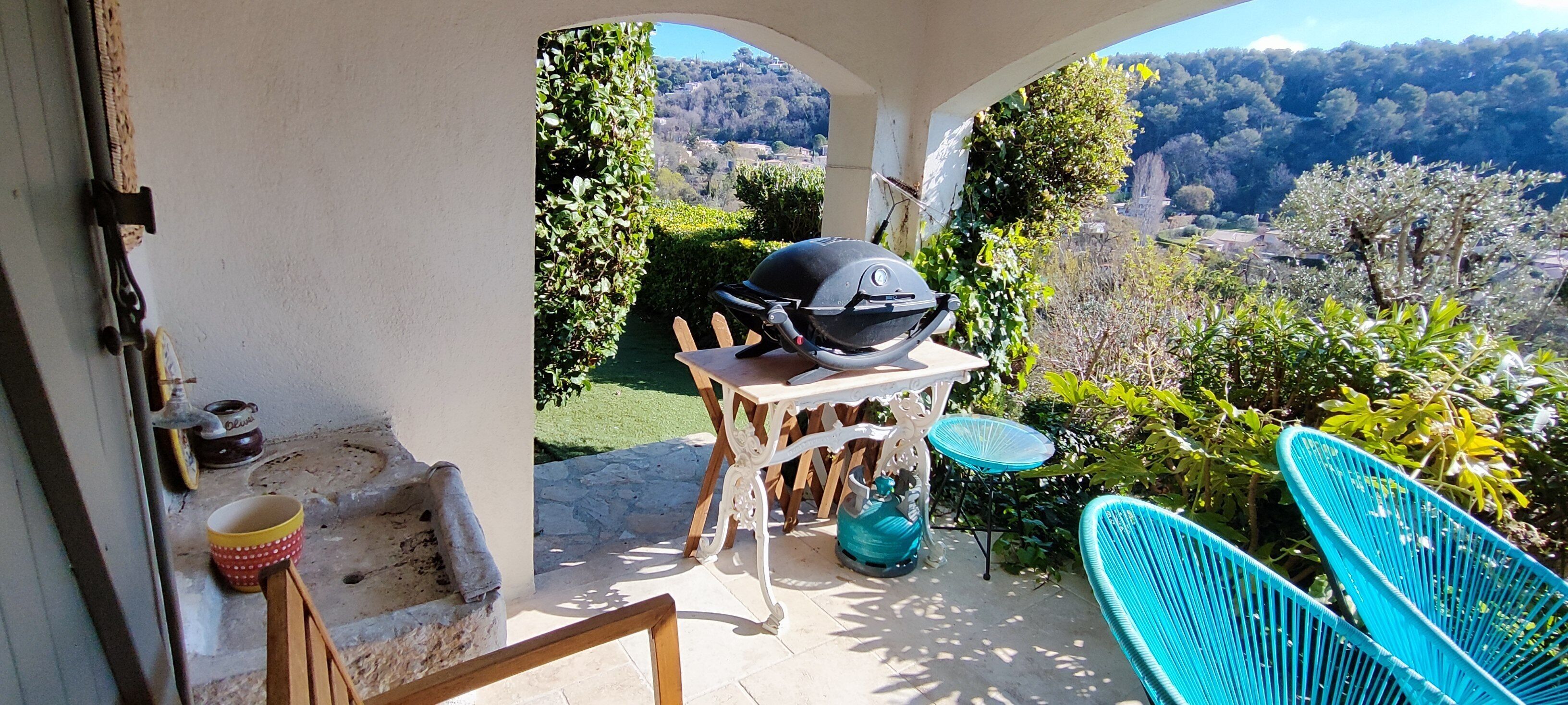A patio with a grill, a table, and chairs, overlooking a garden and a hilly landscape.