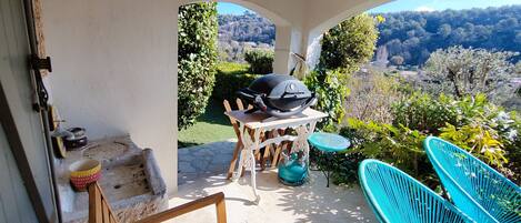 A patio with a grill, a table, and chairs, overlooking a garden and a hilly landscape.