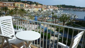 Een balkon met witte stoelen en een klein tafeltje, met uitzicht op een jachthaven met boten en kleurrijke gebouwen op de achtergrond.