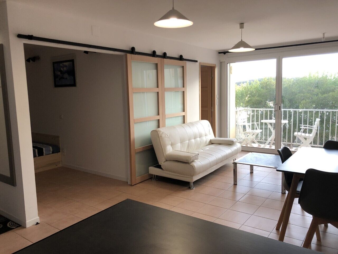 Un salon moderne avec un canapé en cuir blanc, une petite table et un balcon aménagé avec du mobilier d’extérieur.