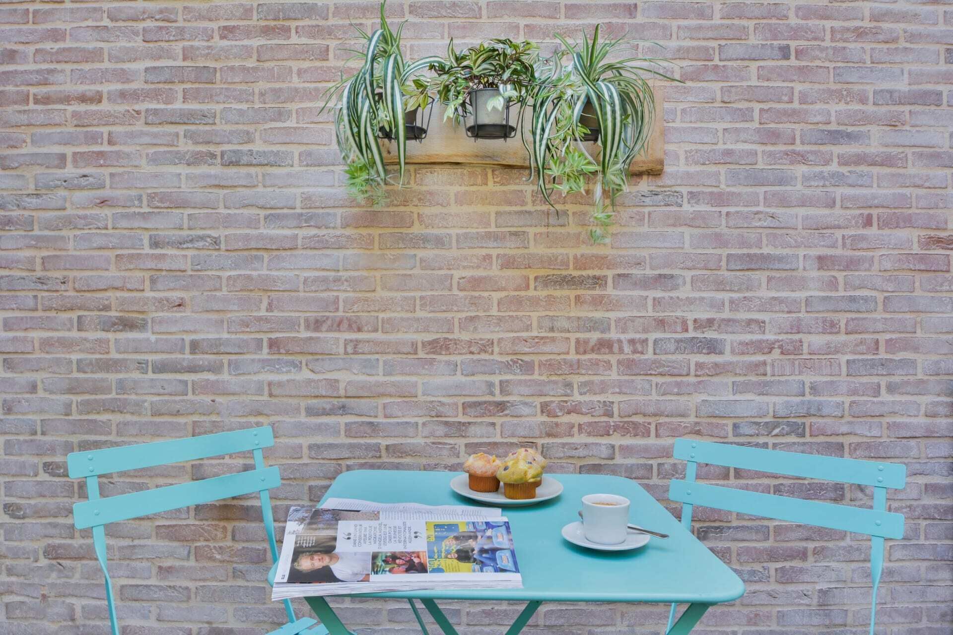 Une table de couleur turquoise, avec un journal, deux muffins et une tasse de café, le tout placé devant un mur de briques orné de plantes suspendues.
